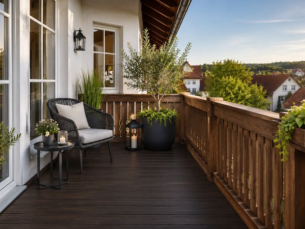 Balkon mit dunklem Holzboden, Holzgeländer, zwei Pflanzen in großen Töpfen, einem kleinen Tisch mit Kerzen und einem Stuhl mit Kissen vor weißen Fenstertüren