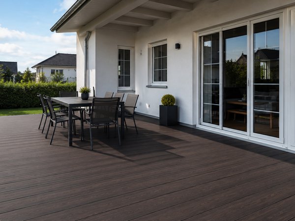 Terrasse mit dunkelbraunem Holzbelag, schwarzem Gartentisch und Stühlen vor einem weißen Haus mit großen Fenstern