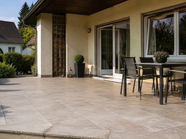 Terrasse mit großformatigen hellen Bodenfliesen, einem Tisch mit vier Stühlen und Pflanzen in Töpfen vor einem Haus mit großen Fenstern